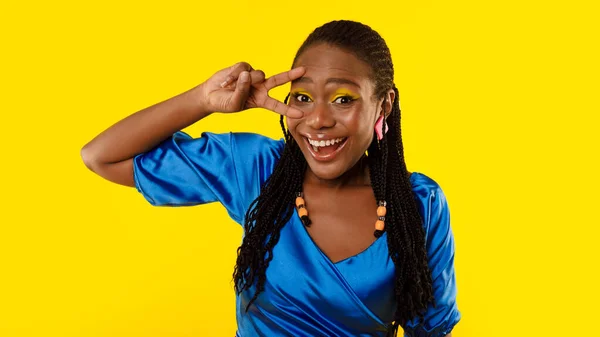 Cheerful Black Woman With Bright Makeup Holding Hand Near Eye Gesturing V Sign Posing In Studio Over Yellow Background, Smiling To Camera. Victory And Peace Concept. Panorama