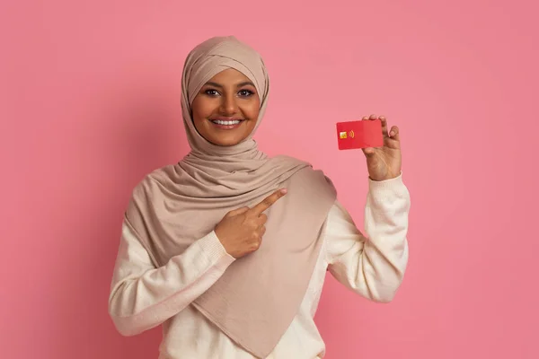 Smiling Muslim Female In Hijab Pointing At Credit Card In Her Hand And Looking At Camera, Happy Middle Eastern Woman Recommending Bank Services While Standing Over Pink Studio Background, Copy Space