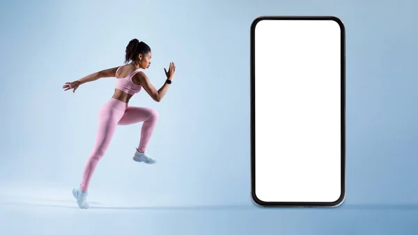 New fitness app. Athletic black woman running, exercising near huge mobile phone over blue background, mockup. Sport motivation and online gym concept