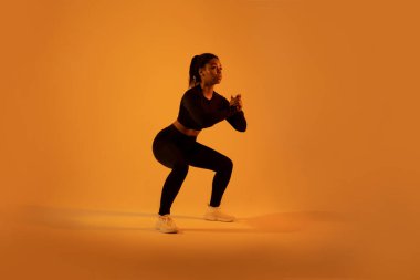 Antrenman ve spor. Sportif siyahi kadın derin çömelme hareketleri yapıyor turuncu neon renkli stüdyo arka planı üzerinde egzersiz yapıyor, tam boy. Spor motivasyonu kavramı