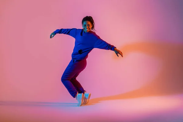 Street dance style. Happy black female hip-hop dancer dancing in neon pink lights, studio background, free space. Sport achievement, spirit of expression
