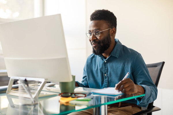 Happy african american man entrepreneur sitting at worktable, looking at computer screen and taking notes at office, looking for new business opportunities, copy space