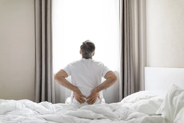 Back view of mature grey-haired man wearing white pajamas sittting on bed at home in the morning, touching lower back, suffering from muscle strain after waking up, copy space