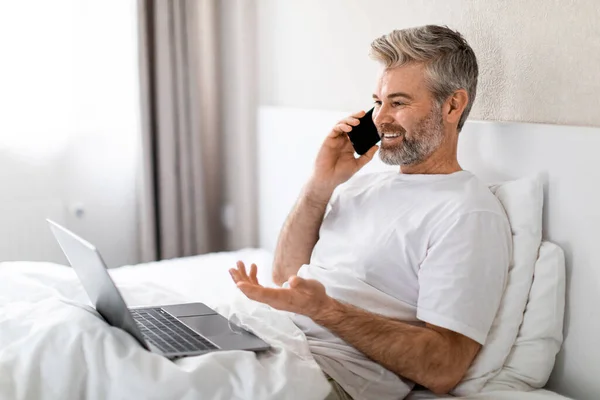 穿着睡衣坐在家里或宾馆床上 在笔记本电脑上工作 与助手或商业伙伴进行电话交谈 复制空间的英俊 白发苍苍的中年商人 — 图库照片