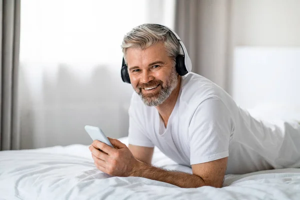 stock image Emotional happy attractive middle aged man wearing pajamas lying on bed at home, holding cell phone, enjoying his brand new headphones, listening to music and smiling at camera, copy space