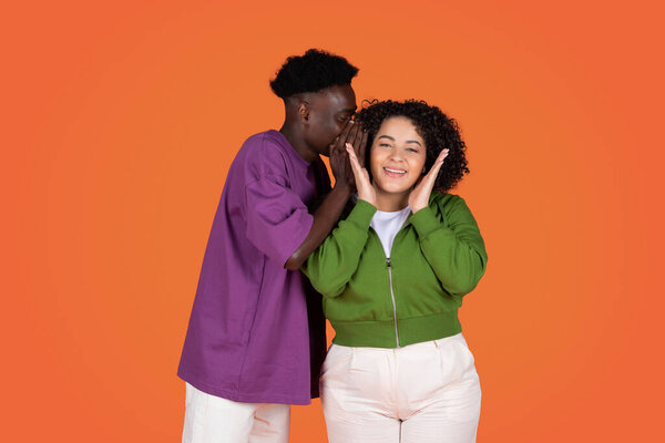 Handsome young african american man sharing secret or whispering gossips into his girlfriends ear, excited obese hispanic woman gesturing, red studio background, copy space