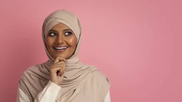 有趣的报价 头戴头巾的好奇的穆斯林女人 把目光投向一边 触摸着下巴 感兴趣的阿拉伯女人头戴头巾 站在粉色工作室的背景上 全景与复制空间 — 图库照片