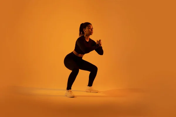 トレーニングとフィットネス オレンジ色のネオンスタジオの背景 全長にわたって行使深いスクワット曲げ筋肉を行うスポーティ黒人女性 スポーツモチベーションコンセプト — ストック写真