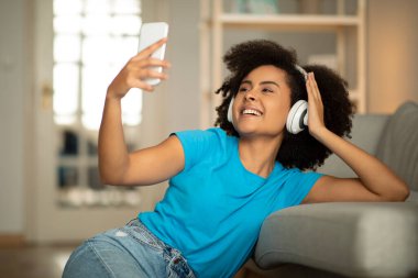 Bin yıllık neşeli, kablosuz kulaklıklı siyah kıvırcık kadın yerde oturmuş akıllı telefonuyla müzik dinliyor Selfie 'nin oturma odasında boş vakit geçirmesini sağlıyor. Eğlence ve eğlence, ses uygulaması