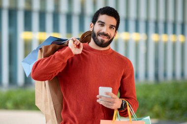 Mobil uygulamayla akıllı telefon alışverişi yapan mutlu Orta Doğulu adam dışarıda kağıt torbalarla kamera pozu verirken gülümsüyor. Alışveriş Başvurusu Reklam Kavramı