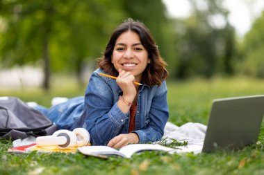 Laptop ve kitap okuyan neşeli genç Arap kız öğrenci şehir parkında çimlerde uzanan, elinde kalem tutan ve kameraya gülümseyen mutlu bir Orta Doğulu kadın.