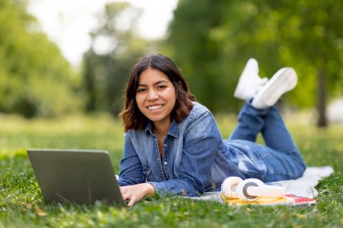 Gülümseyen Arap kız öğrenci dışarıda dizüstü bilgisayar ve çalışma kitaplarıyla çalışıyor, genç Ortadoğulu serbest çalışan kadın bahçede çimlerde uzanmış kameralara, boş alanlara bakıyor.