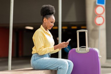 Havalimanı yakınındaki bavulla oturan, akıllı telefon kullanan hoşnutsuz siyah kadın turist, mobil uygulamayla ilgili bir sorun yaşıyor. Seyahat ve İletişim Konsepti