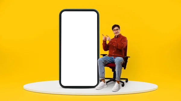巨大な携帯電話で指差し指を指している眼鏡で千年の白人男性を笑顔 広告をお勧めしますし 黄色のスタジオの背景にステージ上の椅子に座る パノラマ — ストック写真