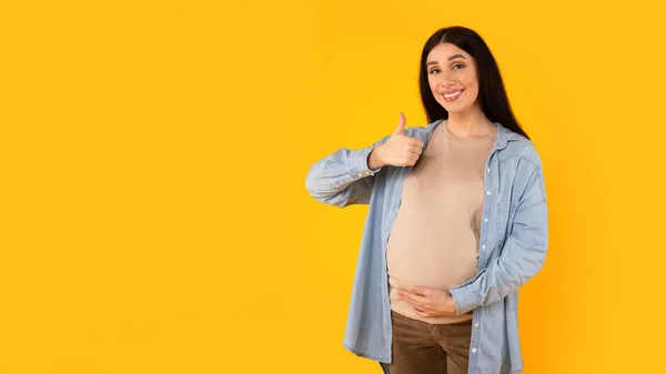 兴奋的孕妇摸着肚子 用手做积极的手势 竖起大拇指 认可某事 站在黄色的背景上 全景尽收眼底 — 图库照片