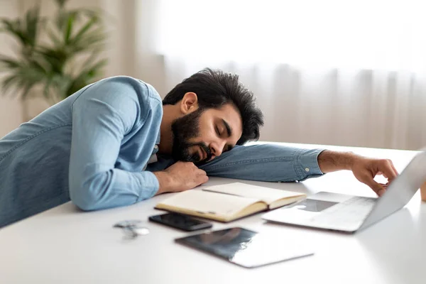 Tired indian male freelancer sleeping at desk in home office, young eastern man leaning on table, napping at workplace, feeling exhausted after working on laptop computer, copy space