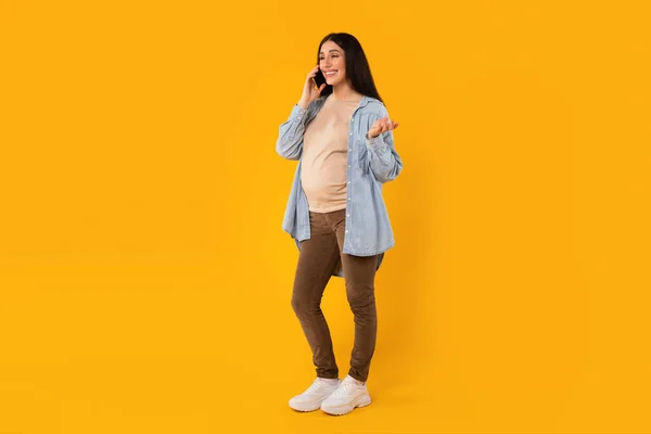 快乐的年轻孕妇一边站在黄色工作室的背景上一边用手机聊天 一边拍摄全长镜头 自由自在 期待着女人和她的医生交谈 — 图库照片