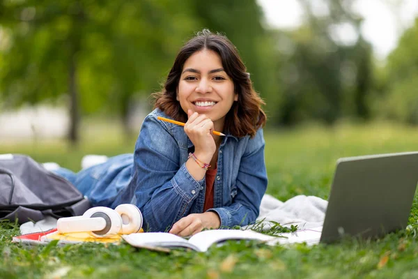 快乐的阿拉伯女学生 带着笔记本电脑和书本在户外学习 快乐的中东千年女性躺在城市公园的草坪上 拿着铅笔 在相机前微笑 闭门造车 — 图库照片