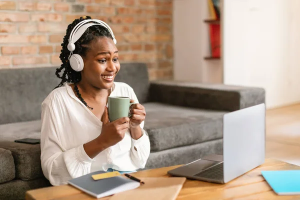 興奮した黒人女性はコーヒーを飲むと自宅でノートパソコンで作業し 女性はヘッドフォンを着用し 画面を見て 黒人女性研究オンラインと笑顔 — ストック写真