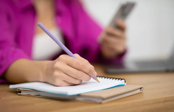 Closeup shot of woman holding smartphone and writing in notepad, unrecognizable female sitting at desk in office, managing plans or making to-do list, noting information to notebook, cropped