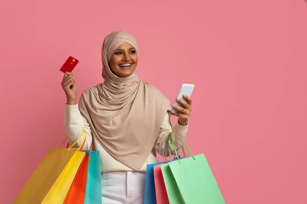 带着智能手机 读卡卡和购物袋的伊斯兰女士微笑站在粉红工作室背景 复制空间旁 头戴头巾在网上购物的快乐穆斯林妇女 — 图库照片