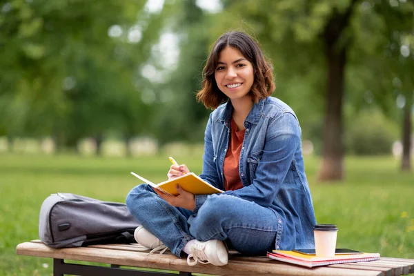 坐在户外长椅上 笑着用笔记本写东西的年轻的中东女人 把快乐的阿拉伯女学生的肖像记在笔记本上 准备在大学校园外参加考试 — 图库照片