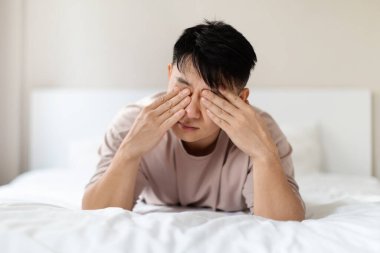 Uykulu, yorgun, Asyalı bir adamın evde pijama giyip, sabah uyandıktan sonra gözlerini ovuşturduğu, kötü uyuduğu, güçsüz hissettiği yakın plan fotoğrafı. Uykusuzluk, uyku bozukluğu konsepti