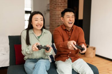 Oturma odasındaki koltukta oturan, elinde joysticklerle gülen, evde video oyunu oynayan, hafta sonlarını birlikte geçiren havalı, mutlu Asyalı eşler.