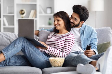 Rahatlamış, neşeli, genç, çok kültürlü eşler hafta sonu evdeki kanepede birlikte takılıp dizüstü bilgisayara bakıyor ve gülümsüyor, patlamış mısır yiyor, Arap adam uzaktan kumanda tutuyor.