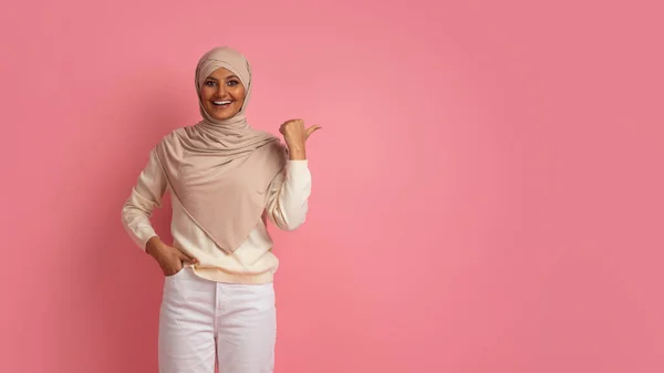 美丽的穆斯林妇女带着她的大拇指在复印空间旁边指指点点 在希吉布微笑伊斯兰女性展示广告的自由空间 同时在粉红工作室的背景下张贴 — 图库照片