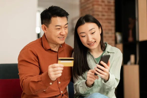快乐的亚洲丈夫和妻子带着智能手机和信用卡在网上购物 坐在客厅的沙发上 现代小玩艺儿和应用程序 购物狂们一起享受销售 — 图库照片