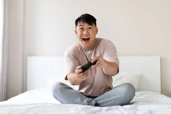 在家里休息 家庭娱乐的概念 快乐的中国成年人坐在床上 手握操纵杆大喊 独自玩电子游戏 复制空间 — 图库照片