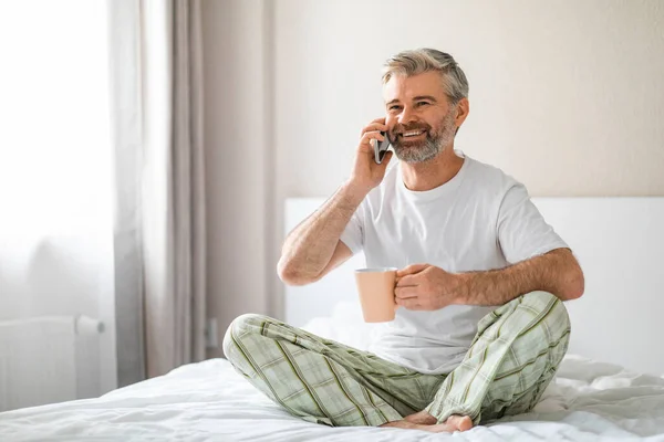 快乐快乐的微笑迷人的中年白发苍苍的男人坐在床上 喝咖啡 打电话 看广告版面 穿睡衣 — 图库照片