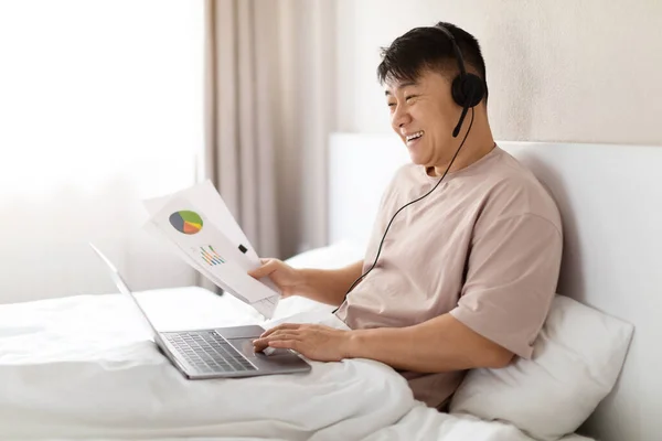 Remote job. Happy handsome middle aged asian man in pajamas manager sitting on bed at home, have video call with team, using computer and headset, reading papers, copy space, side view