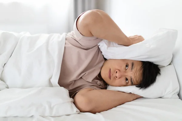 Portrait of sleepless nervous middle aged asian man lying in bed and covering ears with pillow, hearing and suffering from too loud sound or snoring, tired of noisy neighbors. copy space