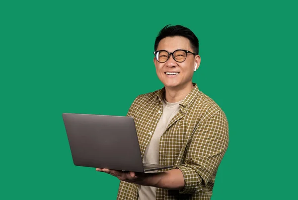Pozitivní Asiat Drží Notebook Usmívá Kameru Sluchátka Brýle Pózuje Zeleném — Stock fotografie
