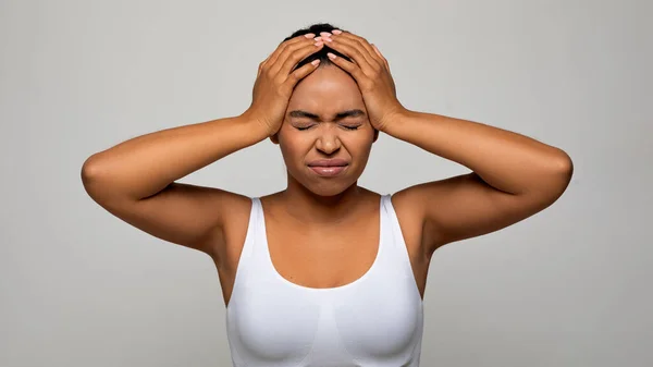 Unhappy sick young african american woman suffer from terrible strong head pain, touching her head with closed eyes, studio shot, black lady have migraine, grey background, copy space