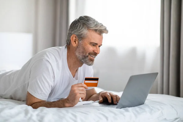 E-commerce, retail. Cheerful handsome middle aged man with beard chilling on bed, using modern pc laptop and plastic bank card, shopping on Internet while staying home at weekend, copy space