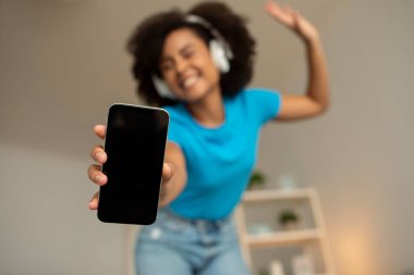 Kablosuz kulaklıklı, gülümseyen siyah kıvırcık kadın müzik dinliyor, boş ekranlı akıllı telefonu gösteriyor ve oturma odasının içinde boş vakit geçiriyor. Eğlence ve eğlence için web sitesi, ses uygulaması