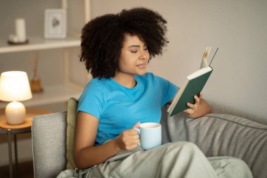 Bin yıllık Afro-Amerikan kıvırcık kadın kitap okumasına sevindim. Oturma odasındaki kanepede dinlenip çay içmenin tadını çıkarıyorum. Kahve molası, akşamları hobi, dinlenme ve ders çalışma, günlük.