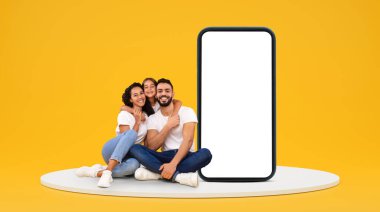 Bin yıllık mutlu Arap ebeveynler ve küçük kız kucaklaşmaları sarı arka planda, stüdyoda boş ekranlı kocaman bir telefonla yerde otururlar. Aile uygulaması iletişimi, web sitesi ve blog