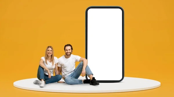 令人欣喜的是 穿着白色T恤的一千年欧洲男人和女人坐在地板上 带着巨大的空白屏幕的电话 隔离在黄色背景 工作室和复制空间中 应用程序 广告和好的报价 — 图库照片