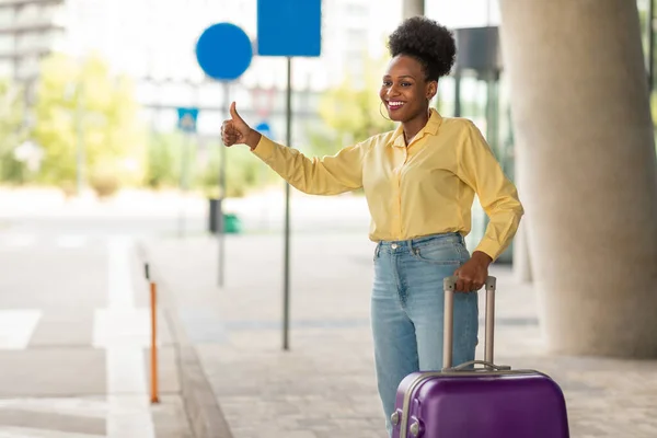 Taxi Ride. Smiling African American Female With A Suitcase Showing Sign Catching A Cab Standing Outdoors In Urban Area Near Airport. Tourism And City Transportation Service Concept