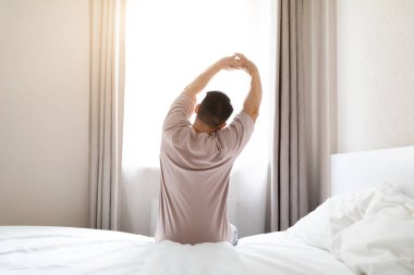 Esmer adamın pijama giydiği ve sabah uyandıktan sonra vücudu gerdiği pencereye bakıp uzayı kopyaladığı arka plan görüntüsü. Rahat, sağlıklı uyku konsepti