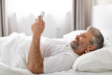 Rahatlamış, mutlu, beyaz tişörtlü, beyaz sakallı olgun bir adam sabah uyandıktan sonra yatakta dinleniyor, akıllı telefon kullanıyor ve gülümsüyor, sosyal medyada kayıyor.
