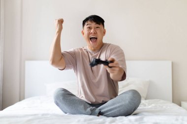 Mutlu, yakışıklı, orta yaşlı Asyalı oyuncu evde eğleniyor, yatakta oturuyor, joystick tutuyor ve elini kaldırıyor zaferi kutluyor, video oyunu oynuyor, pijama giyiyor, fotokopi çekiyor.