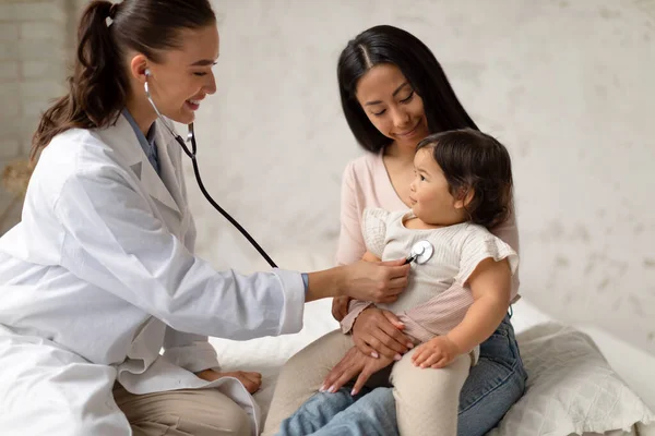 Happy Female Doctor Doing Baby Health Checkup Listening To Japanese Little Girls Heart Via Stethoscope As Cute Toddler Sitting With Mommy In Modern Clinic. Family Healthcare Concept