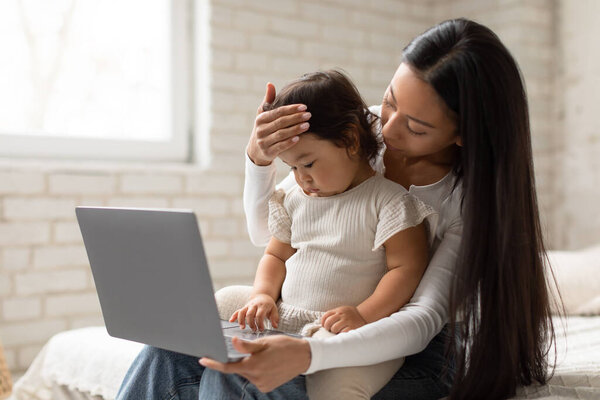 Japanese Mom And Baby Girl Using Laptop Having Online Medical Appointment At Home. Mother Video Calling Doctor Sitting With Sick Toddler Daughter In Bedroom, Touching Forehead. Childhood Diseases