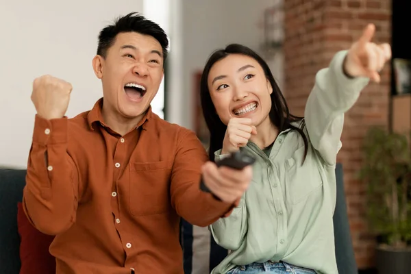 Joyful asian spouses shaking fists and shouting, watching sport channel, woman pointing on TV, sitting on sofa at home. Sport and television programming, weekend leisure