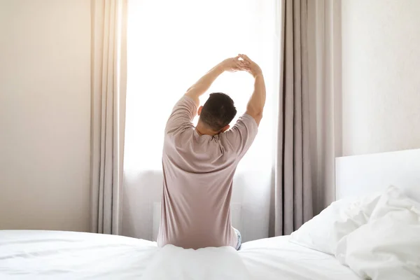 朝目が覚めた後 ベッドの上に座って体を伸ばしパジャマを着たブルネットの男のバックビューは ウィンドウを見て スペースをコピーします 快適な健康的な睡眠コンセプト — ストック写真